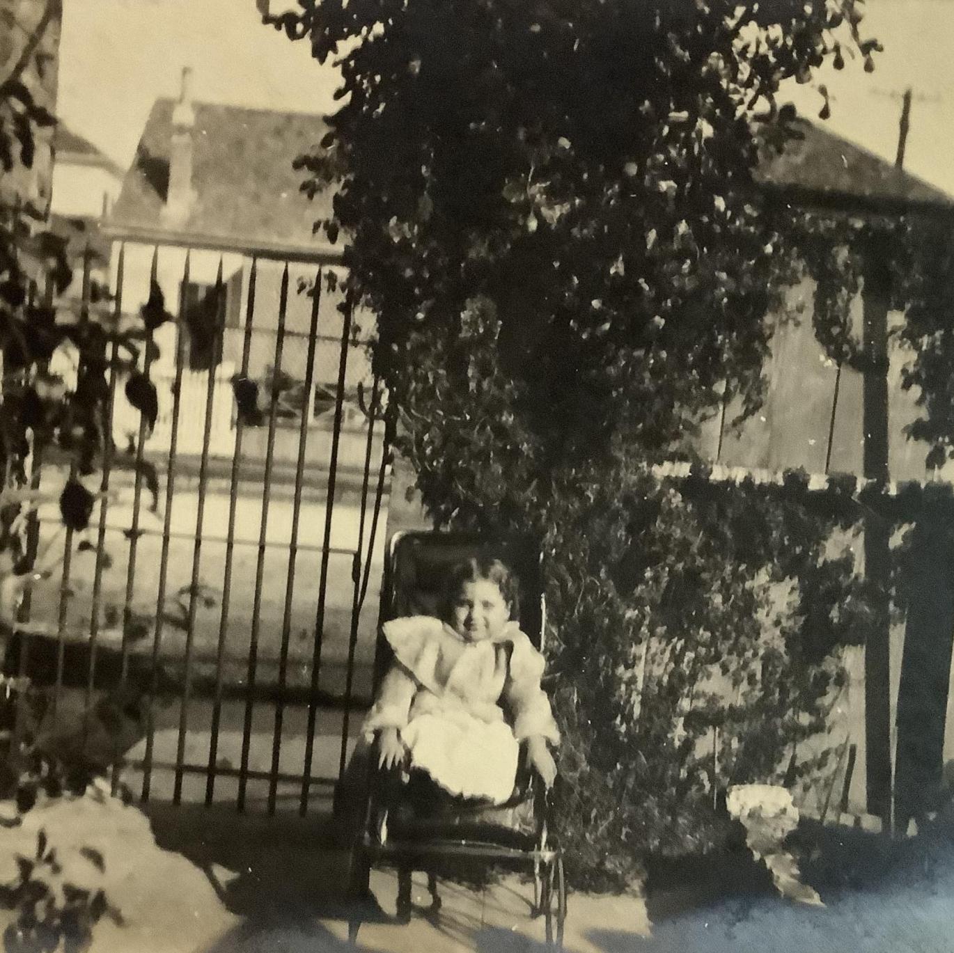 A yellowed black and white photo of a girl in a frilly dress sitting in a wheelchair. Sheehan posed in front of a large house with vines growing decoratively over the gate