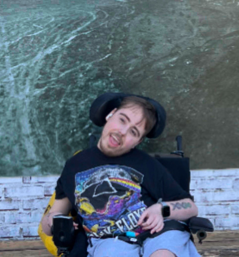 A young man in a powered wheelchair poses in front of a mural on a brick wall. He is white with short brown hair and a beard. He is wearing a Pink Floyd shirt and has a tattoo of DNA on his left arm.